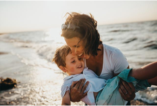 Photograph of a mother holding her son in her arms on the beach, both of them smiling.